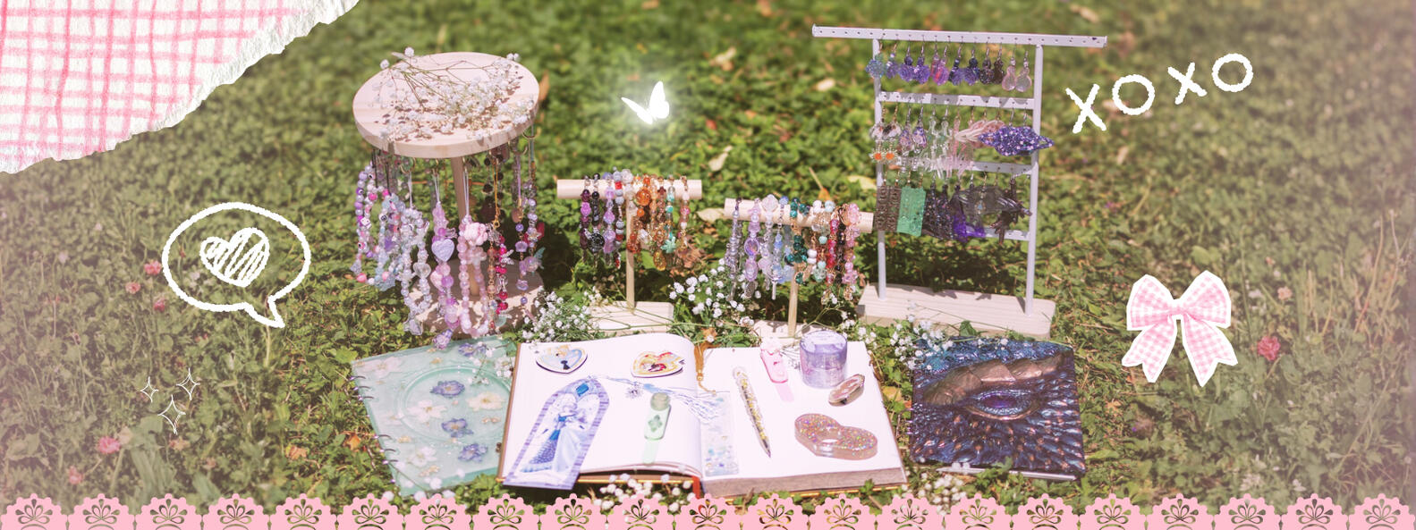 Accessoires de lecture, mode et décoration - Yuri dessine fantaisie Divers présentoirs posés sur de l'herbe avec des produits (bracelets de téléphone, bijoux de téléphone, porte clés, bracelets astrologiques, boucles d'oreilles, etc) et devant un livre ouvert ainsi que deux livres en résine fermés d'un côté et d'autre, ave